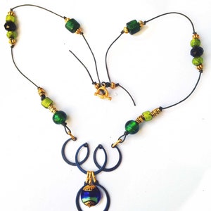 May include: A handcrafted necklace featuring a dark blue metal pendant with a blue and green glass bead. The necklace has green and gold beads strung on a black cord, with a gold clasp. The design is artistic and unique.