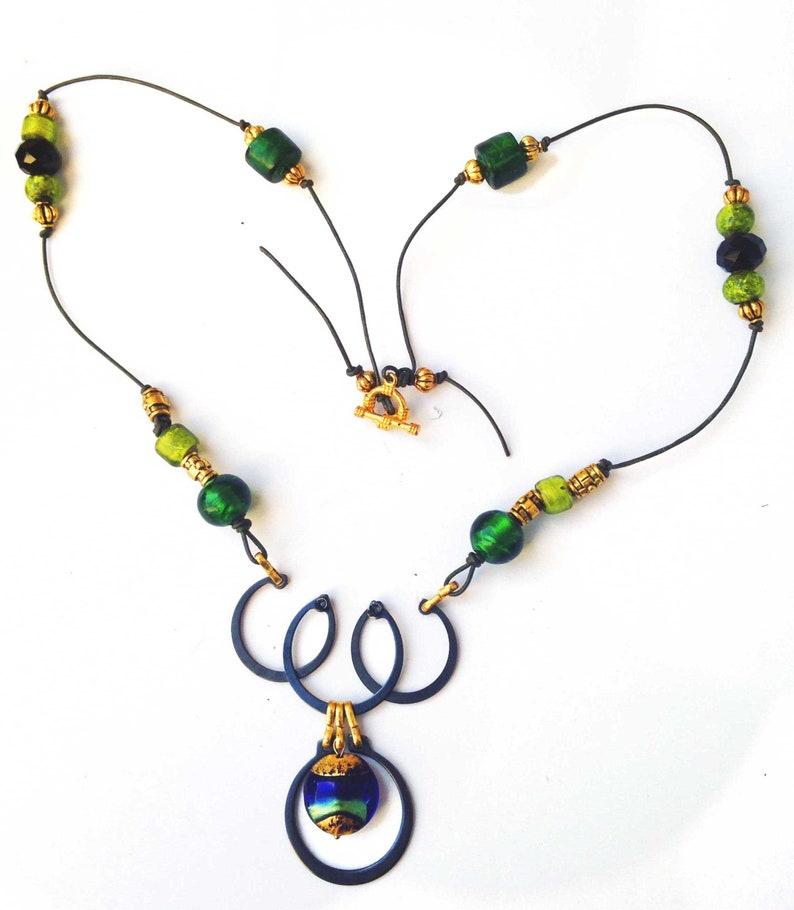 May include: A handcrafted necklace featuring a dark blue metal pendant with a blue and green glass bead. The necklace has green and gold beads strung on a black cord, with a gold clasp. The design is artistic and unique.