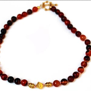 May include: A necklace featuring a strand of reddish-brown beads with a gold-colored clasp. The beads vary in color, with some appearing darker than others. The necklace also includes gold-colored decorative elements.