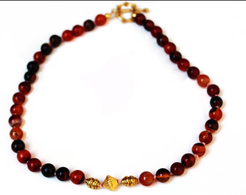 May include: A necklace featuring a strand of reddish-brown beads with a gold-colored clasp. The beads vary in color, with some appearing darker than others. The necklace also includes gold-colored decorative elements.