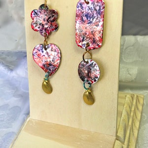 May include: Artistic dangle earrings with a floral, heart, and rectangular design. The earrings feature a colorful abstract pattern in red, purple, and white. Each earring has a gold-colored charm at the bottom and is connected with gold-colored rings.