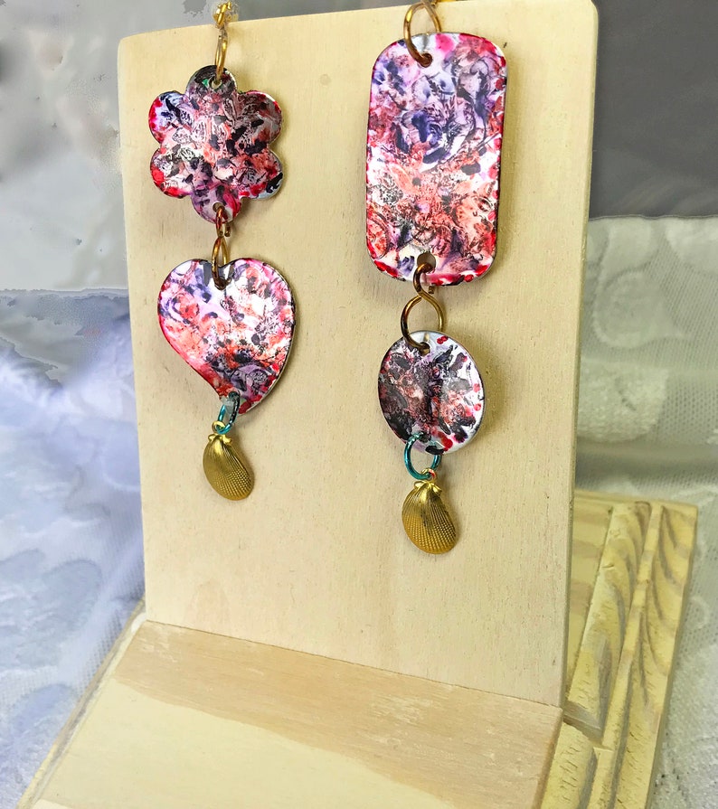 May include: Artistic dangle earrings with a floral, heart, and rectangular design. The earrings feature a colorful abstract pattern in red, purple, and white. Each earring has a gold-colored charm at the bottom and is connected with gold-colored rings.