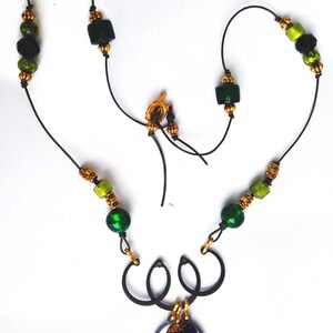 May include: A handcrafted necklace featuring a black cord strung with green and gold beads. The necklace has a gold clasp and a pendant with a dark metal frame and a colorful, iridescent center. The design is intricate and eye-catching.
