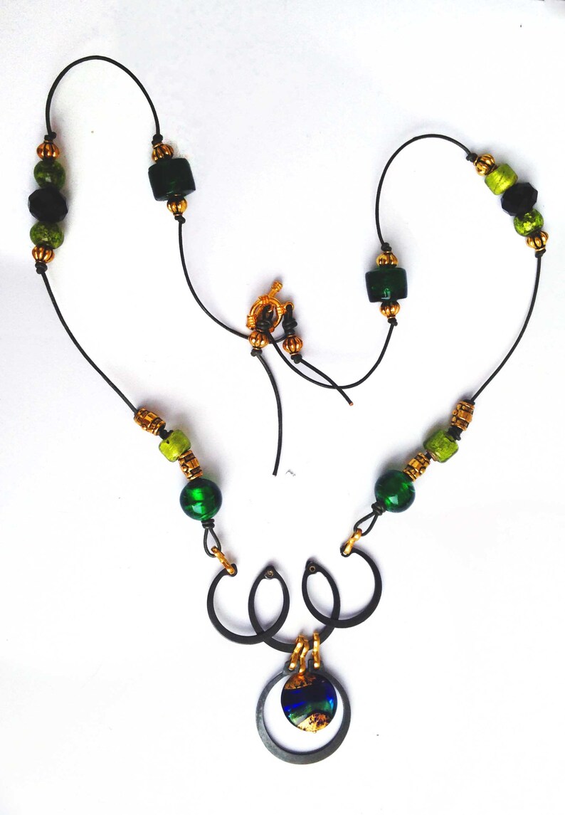 May include: A handcrafted necklace featuring a black cord strung with green and gold beads. The necklace has a gold clasp and a pendant with a dark metal frame and a colorful, iridescent center. The design is intricate and eye-catching.