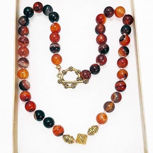 May include: A beaded necklace featuring a variety of earth-toned beads in shades of red, orange, and black. The necklace has a gold-colored clasp and decorative beads. The necklace is presented in a white box with a gold-colored rim.