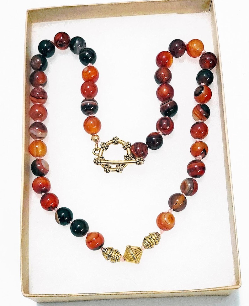 May include: A beaded necklace featuring a variety of earth-toned beads in shades of red, orange, and black. The necklace has a gold-colored clasp and decorative beads. The necklace is presented in a white box with a gold-colored rim.