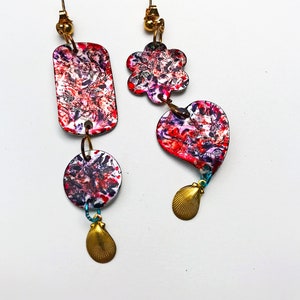 May include: Artistic dangle earrings with a unique design. One earring features a rectangular shape, a circle, and a gold seashell charm. The other earring has a flower and heart shape with a gold seashell charm. Both earrings have a colorful, abstract pattern.