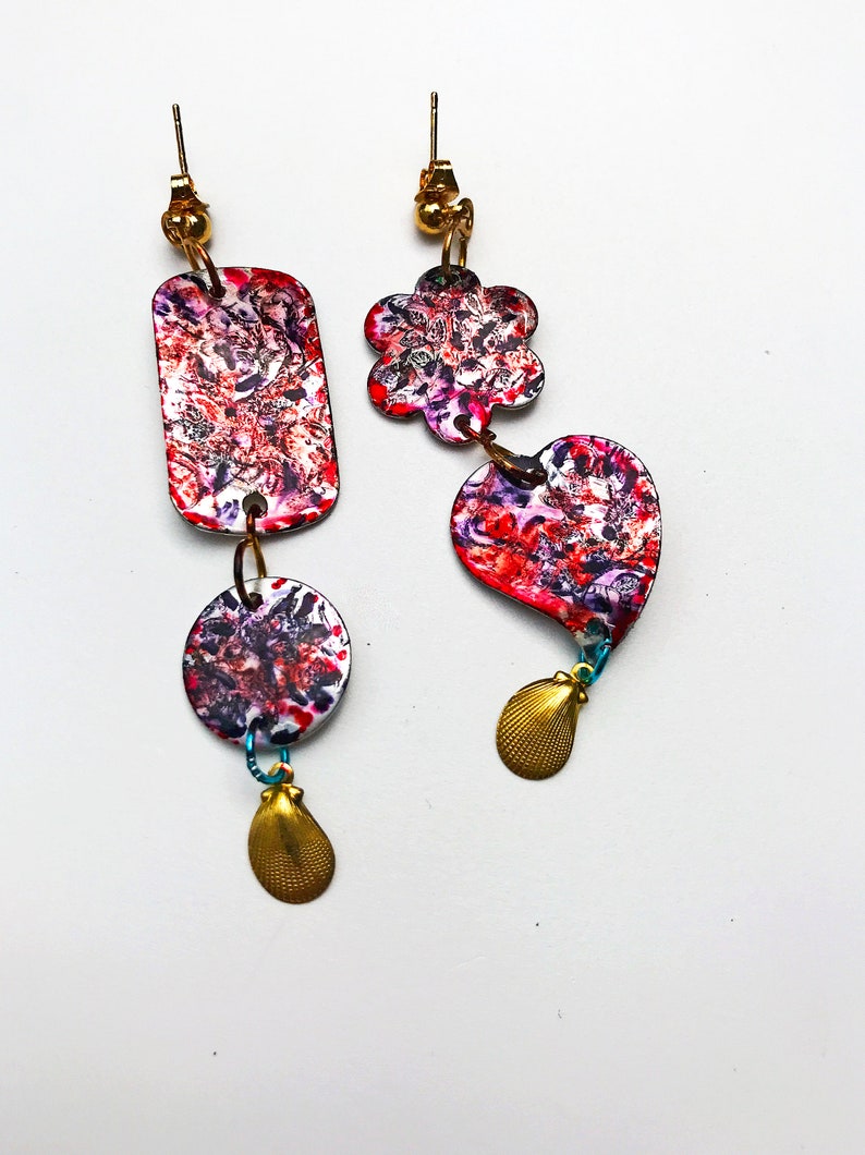 May include: Artistic dangle earrings with a unique design. One earring features a rectangular shape, a circle, and a gold seashell charm. The other earring has a flower and heart shape with a gold seashell charm. Both earrings have a colorful, abstract pattern.