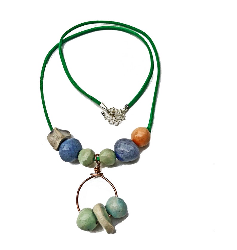 May include: A handmade necklace featuring a green cord and a variety of colorful beads. The beads include shades of blue, green, peach, and gray. A copper wire circle holds additional beads, creating a unique pendant.