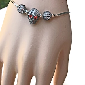 Op de afbeelding: Een zwarte schedelarmband met rode ogen en sprankelende accenten. De schedel is gecentreerd tussen twee kralenbollen, alles op een dunne, zilverkleurige band. De armband is ontworpen om om de pols te worden gedragen.