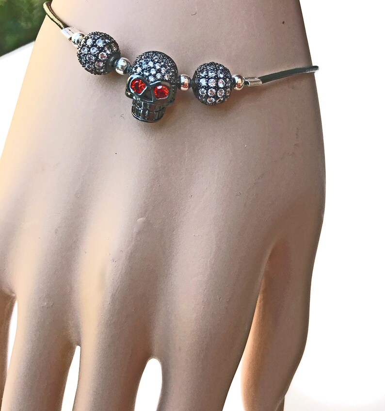 Op de afbeelding: Een zwarte schedelarmband met rode ogen en sprankelende accenten. De schedel is gecentreerd tussen twee kralenbollen, alles op een dunne, zilverkleurige band. De armband is ontworpen om om de pols te worden gedragen.