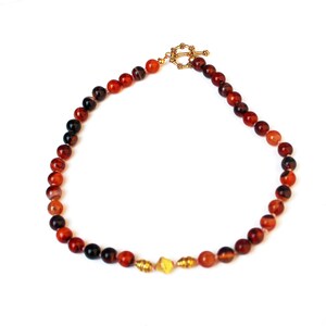 May include: A beaded necklace featuring red and black agate stones. The necklace has a gold-colored clasp and decorative accents. The beads are round and vary in size, creating a natural and earthy aesthetic. The necklace is displayed on a white background.