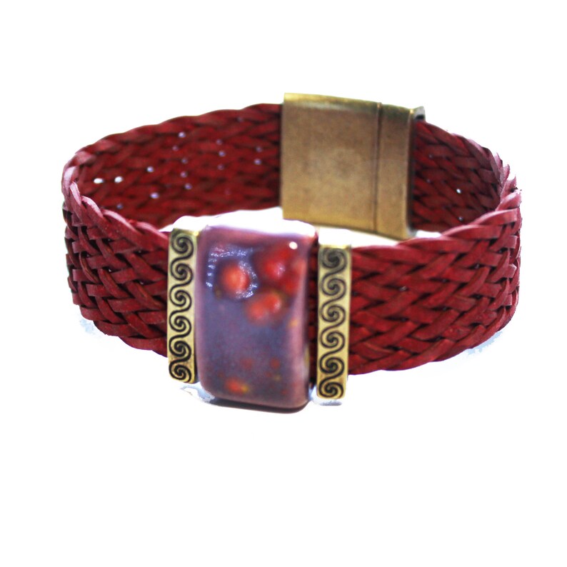May include: A red braided leather bracelet with a rectangular, speckled, purple and red center stone. The stone is framed by gold-colored metal with a decorative swirl design. The bracelet has a gold-colored magnetic clasp.
