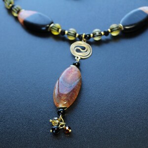 May include: A handcrafted necklace featuring a large, oval pendant with brown and red tones, suspended from a beaded chain. The chain incorporates black and yellow beads, with a gold-colored spiral accent. The necklace is set against a dark background.