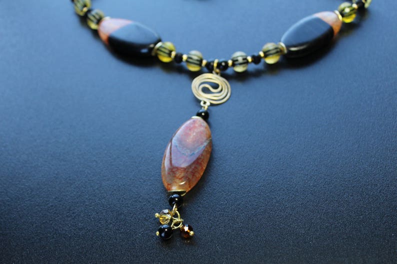 May include: A handcrafted necklace featuring a large, oval pendant with brown and red tones, suspended from a beaded chain. The chain incorporates black and yellow beads, with a gold-colored spiral accent. The necklace is set against a dark background.