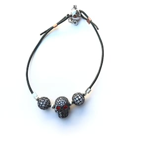 Op de afbeelding: Een zwart leren armband met een schedelhangertje met rode ogen en twee ronde, met kristallen bezette kralen. De armband heeft een magnetische sluiting en zilverkleurige accenten. De schedelhangertje is het middelpunt van dit unieke sieraad.