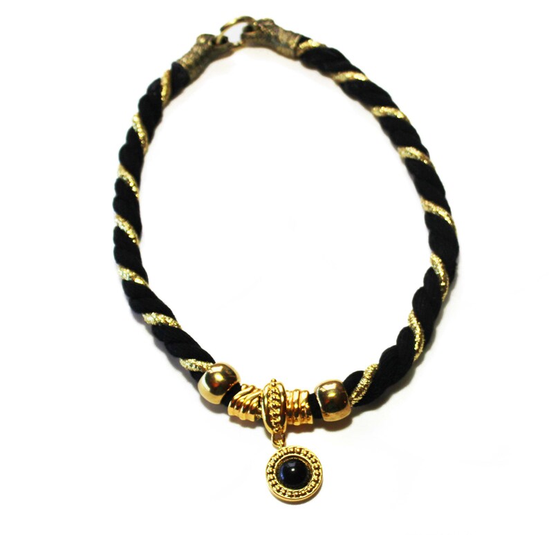 May include: A black and gold rope necklace with a gold clasp. The necklace features a gold pendant with a black stone. The rope is twisted with gold thread.