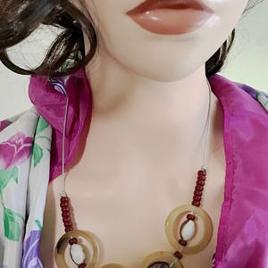 May include: A necklace featuring a silver-toned cord, small dark red beads, and three large translucent tan rings. Each ring contains a central round bead with a black and white swirl pattern, while the top ring holds a white speckled bead.