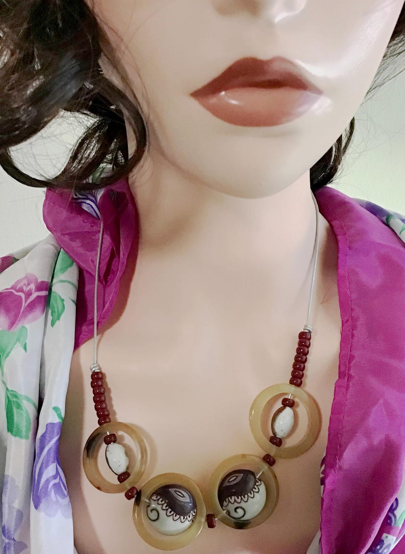 May include: A necklace featuring a silver-toned cord, small dark red beads, and three large translucent tan rings. Each ring contains a central round bead with a black and white swirl pattern, while the top ring holds a white speckled bead.