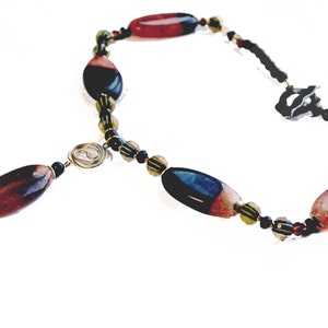 May include: A handcrafted necklace featuring large, oval-shaped beads in shades of red, black, and blue. The necklace includes smaller black and gold beads, with a wire spiral accent and a delicate chain.