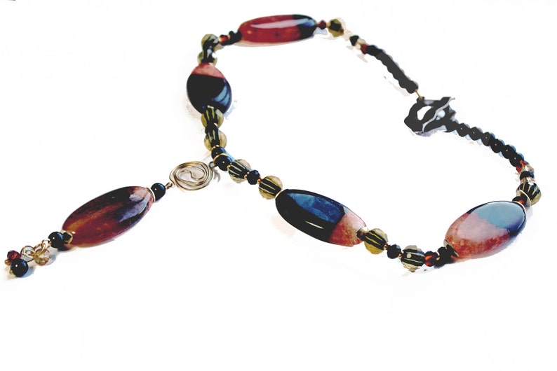 May include: A handcrafted necklace featuring large, oval-shaped beads in shades of red, black, and blue. The necklace includes smaller black and gold beads, with a wire spiral accent and a delicate chain.