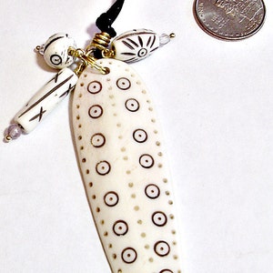 May include: Ivory-colored pendant necklace with a black cord. The pendant is elongated with brown circle and dot patterns. Additional ivory beads with black designs and gold accents hang from the pendant.