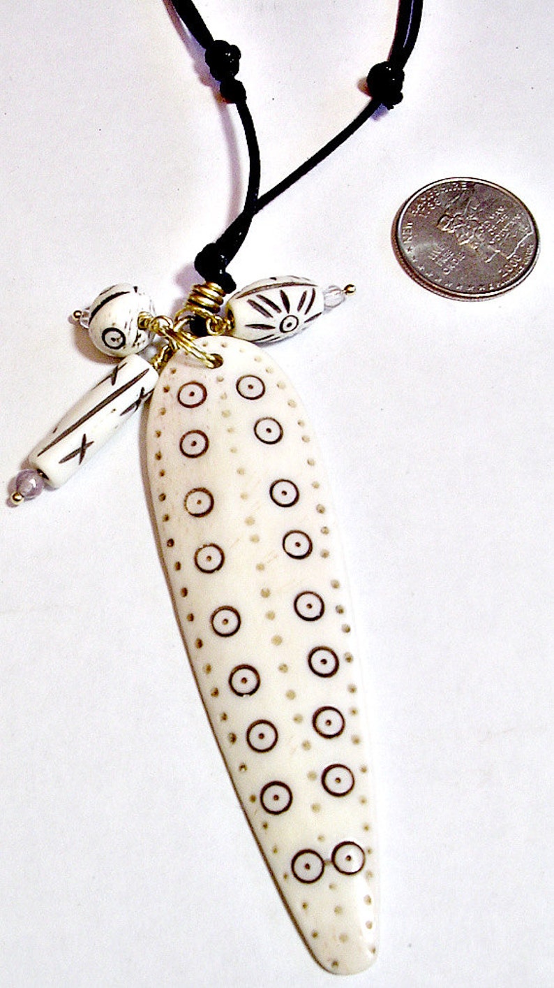 May include: Ivory-colored pendant necklace with a black cord. The pendant is elongated with brown circle and dot patterns. Additional ivory beads with black designs and gold accents hang from the pendant.