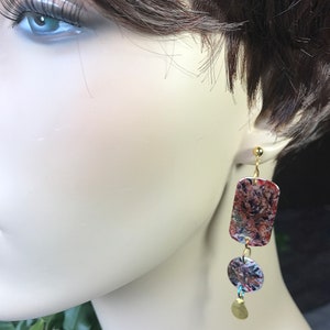 May include: Dangling earrings with a gold-colored post and a rectangular top, a circular middle, and a small gold-colored charm. The earrings have a colorful, abstract pattern in shades of red, brown, and blue.