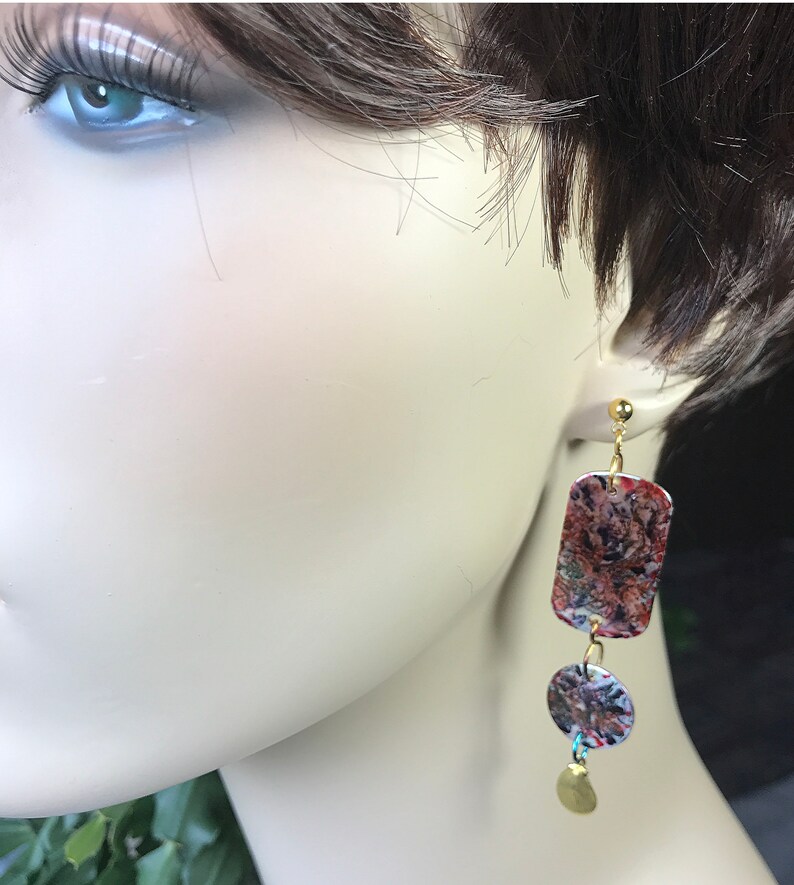 May include: Dangling earrings with a gold-colored post and a rectangular top, a circular middle, and a small gold-colored charm. The earrings have a colorful, abstract pattern in shades of red, brown, and blue.