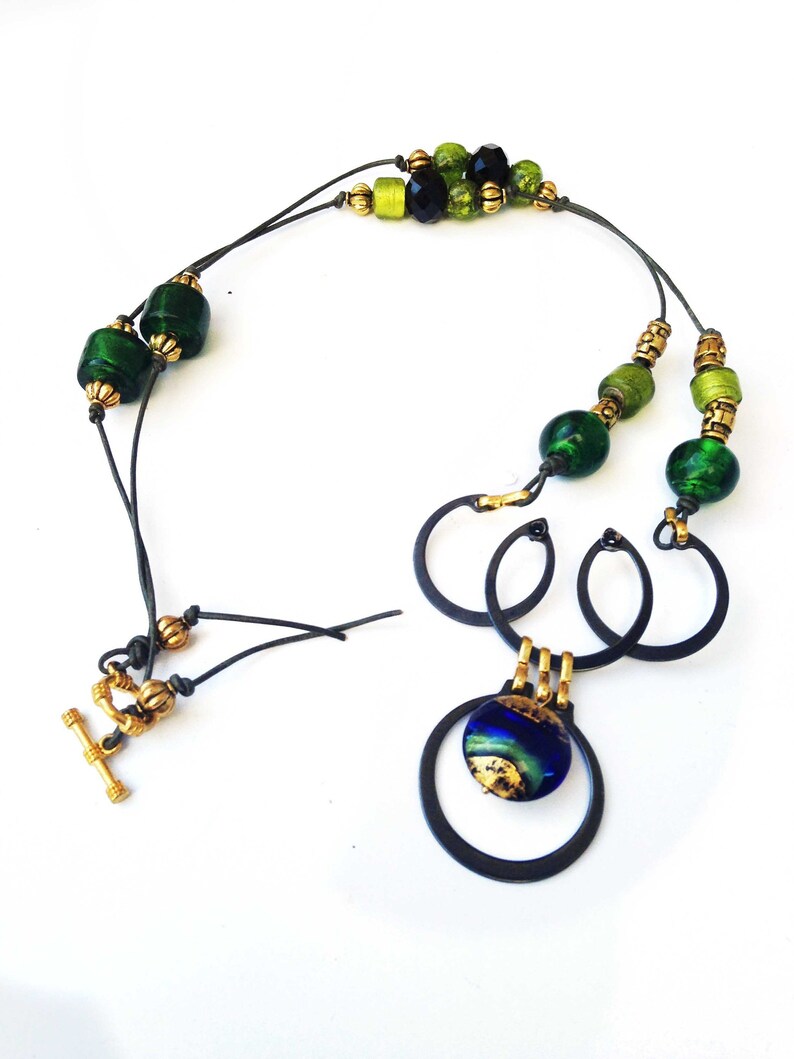 May include: A handcrafted necklace featuring a mix of green and black beads, gold accents, and a unique pendant. The pendant has a blue and gold design, suspended within black metal rings. The necklace has a toggle clasp.