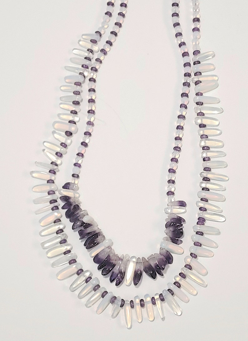 May include: A layered necklace featuring two strands. The top strand has small, clear beads and purple accents. The bottom strand has iridescent, teardrop-shaped beads and a central section of purple, irregular-shaped beads.