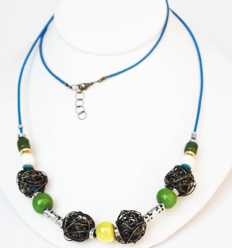 May include: A handcrafted necklace featuring a vibrant blue cord. The necklace showcases a variety of beads, including green, white, and yellow, interspersed with intricate wire cage beads. The design is completed with silver-toned metal accents and a secure clasp.