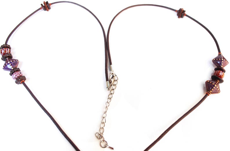 May include: A brown leather necklace with decorative beads. The necklace features square and round beads in shades of purple, red, and brown. The necklace has a silver-colored chain for adjustable length.