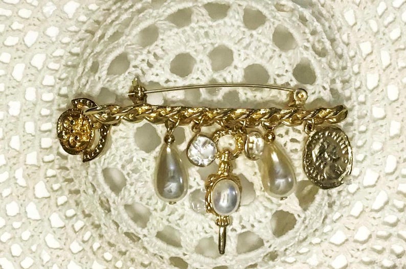 May include: Gold-toned brooch with a safety pin backing. The brooch features a chain-like design with dangling charms, including faux pearls, a coin, and a key-shaped pendant. The brooch is set against a white, lacy background.
