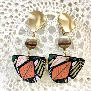 May include: A pair of dangle earrings with gold-colored, irregular-shaped tops. Each earring features a striped bead, a leaf-patterned pendant with orange and white leaves, and gold-colored connecting hardware. The earrings are displayed on a white, open-weave surface.