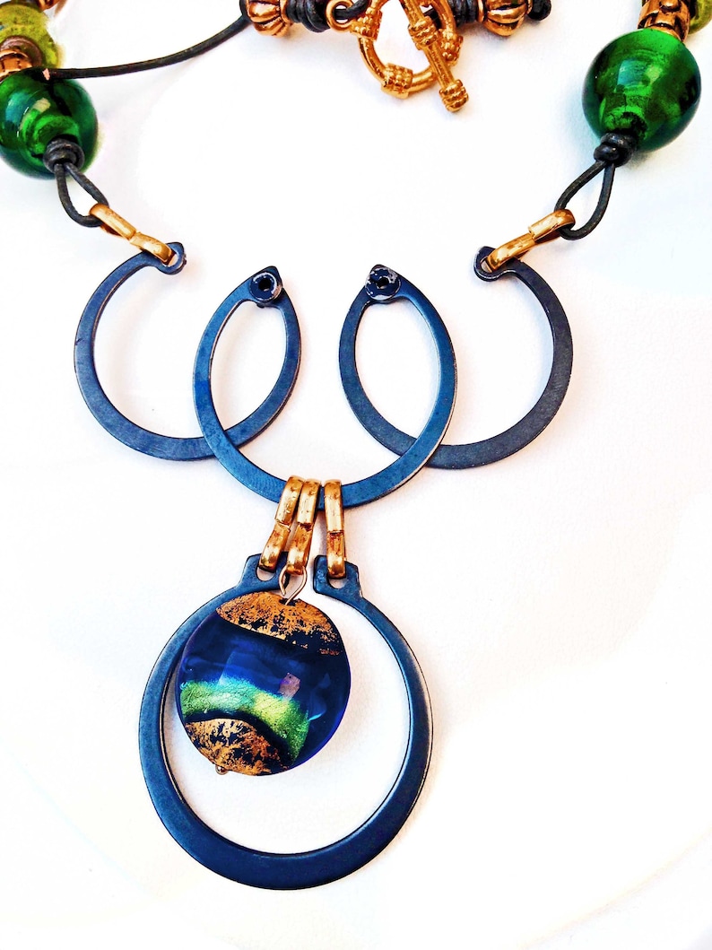 May include: A handcrafted necklace featuring a dark blue metal pendant with a circular design, accented by a dichroic glass bead with gold leaf. The necklace includes green glass beads, leather cord, and gold-toned metal accents. The clasp is a gold-toned toggle.