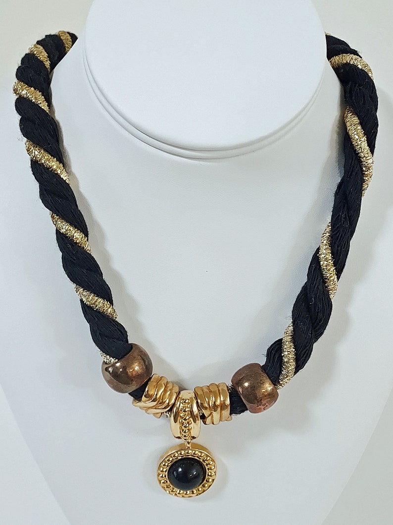 May include: Black and gold braided necklace with a pendant. The necklace features a black braided cord with gold metallic accents. The pendant is a gold-toned circular frame with a black stone center. The necklace has gold and bronze-toned metal accents.