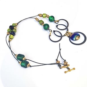 May include: A handcrafted necklace featuring green and gold beads, with a blue and gold pendant. The necklace has a black cord and a gold toggle clasp. The beads vary in size and shade, creating a unique design.