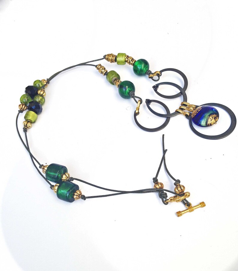 May include: A handcrafted necklace featuring green and gold beads, with a blue and gold pendant. The necklace has a black cord and a gold toggle clasp. The beads vary in size and shade, creating a unique design.