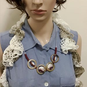 May include: A necklace featuring three circular translucent tan rings, each containing a small white and maroon patterned disc. The necklace is accented with small maroon beads on a silver-toned wire, worn over a blue collared shirt and a ruffled scarf.