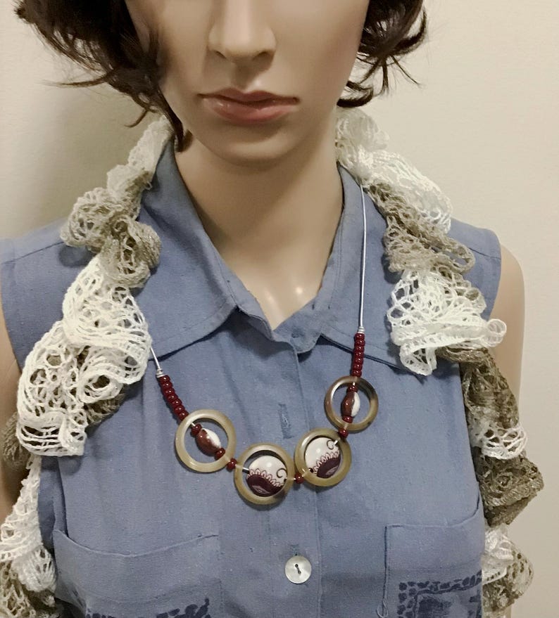 May include: A necklace featuring three circular translucent tan rings, each containing a small white and maroon patterned disc. The necklace is accented with small maroon beads on a silver-toned wire, worn over a blue collared shirt and a ruffled scarf.