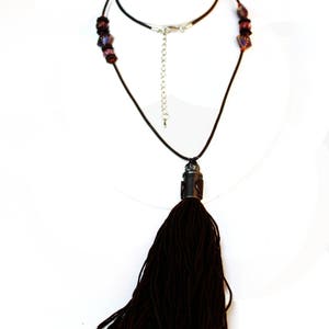 May include: A dark brown tassel necklace with a black cord. The necklace features a decorative metal piece at the top of the tassel and dark red beads near the clasp. The necklace is displayed on a white background.