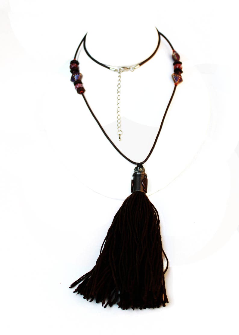 May include: A dark brown tassel necklace with a black cord. The necklace features a decorative metal piece at the top of the tassel and dark red beads near the clasp. The necklace is displayed on a white background.