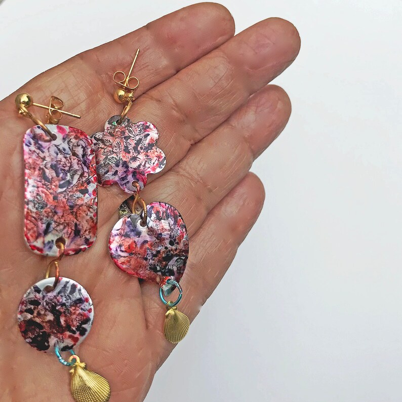 May include: A pair of asymmetrical earrings with a colorful, abstract pattern in shades of red, pink, and purple. The earrings feature various shapes, including a rectangle, a flower, and a circle, with small gold seashell charms. The earrings are held in a hand.
