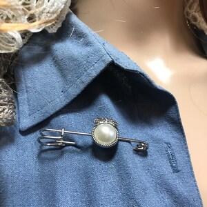 May include: A silver-toned brooch featuring a faux pearl center, surrounded by a twisted rope design, and topped with a small crown. The brooch is pinned to a blue linen shirt.