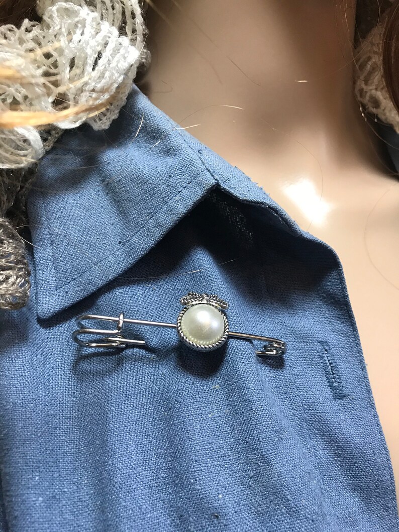 May include: A silver-toned brooch featuring a faux pearl center, surrounded by a twisted rope design, and topped with a small crown. The brooch is pinned to a blue linen shirt.