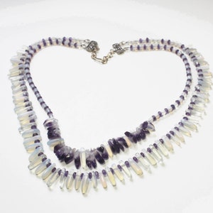 May include: A two-strand necklace featuring a combination of purple and white beads. The necklace has a silver clasp and is composed of small, round beads and larger, teardrop-shaped beads. The design is symmetrical and elegant.