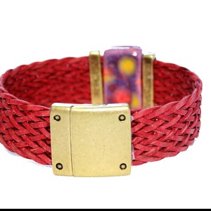 May include: A red woven leather bracelet with a gold-colored magnetic clasp. The bracelet features a rectangular clasp and a decorative rectangular piece with purple, yellow, and white accents. The bracelet is approximately 1 inch wide.