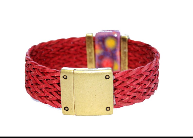 May include: A red woven leather bracelet with a gold-colored magnetic clasp. The bracelet features a rectangular clasp and a decorative rectangular piece with purple, yellow, and white accents. The bracelet is approximately 1 inch wide.