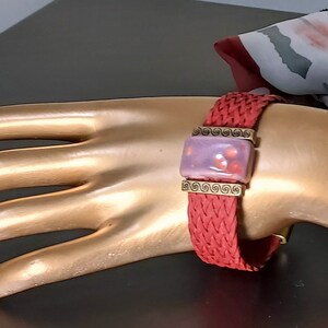 May include: A red braided bracelet with a rectangular lavender and red centerpiece. The centerpiece is framed with gold-colored metal and features a swirl design. The bracelet is displayed on a gold-colored hand.